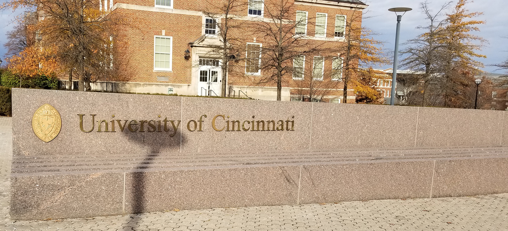 University of Cincinnati sign on a bench in front of a building and trees